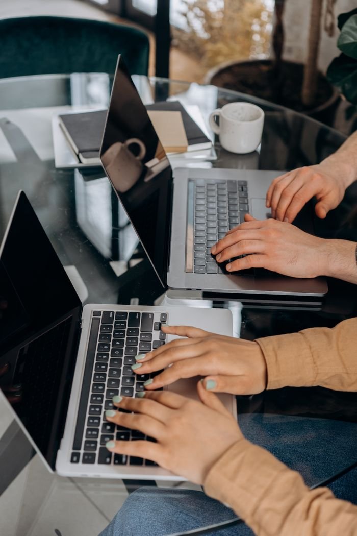 Two adults collaborating remotely on laptops at a glass table indoors.